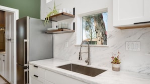A modern kitchen with a stainless steel refrigerator, white cabinetry, a marble countertop, a built-in sink, and a window with a view of trees. - Stunning Guesthouse with Private Entry & Courtyard (Santa Maria)