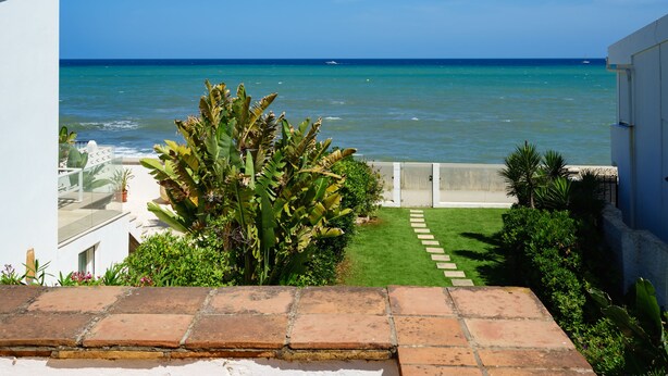 A view of the ocean from a balcony with a tiled floor, a small garden with a palm tree, and a white building with a red-tiled roof.