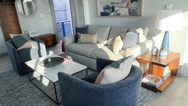 Una sala de estar moderna con un sofá gris, un sillón azul y una mesa de centro. Hay un ventilador de techo, un cuadro en la pared y una mesita de noche con objetos decorativos.