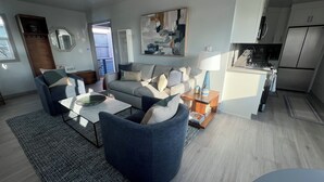 Una sala de estar moderna con sofá, sillones y una mesa de centro. Hay una zona de cocina con refrigerador y microondas.