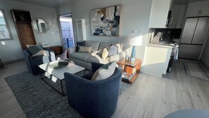 Ein modernes Wohnzimmer mit einer Couch, Sesseln und einem Couchtisch. Es gibt eine Küchenzeile mit einem Kühlschrank und einer Mikrowelle.