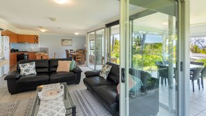 Un salon moderne avec un canapé en cuir noir, une porte-fenêtre en verre donnant sur un balcon et une cuisine en arrière-plan.