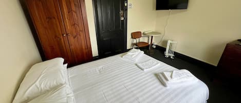 A hotel room with a bed, a wooden wardrobe, a black door, a small table with a chair, and a TV mounted on the wall.