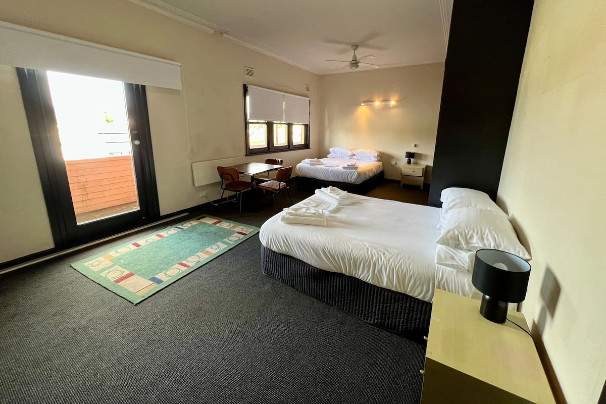 A hotel room with a bed, a desk, a chair, a small table, a nightstand, a lamp, a ceiling fan, and a window with curtains.