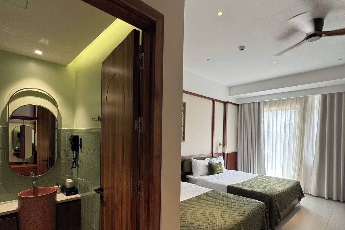 A hotel room with a bathroom, a bed with green bedding, a ceiling fan, and a large window with curtains.