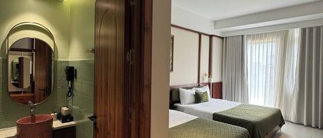Habitación de hotel con baño, cama con ropa de cama verde, ventilador de techo y una ventana grande con cortinas.
