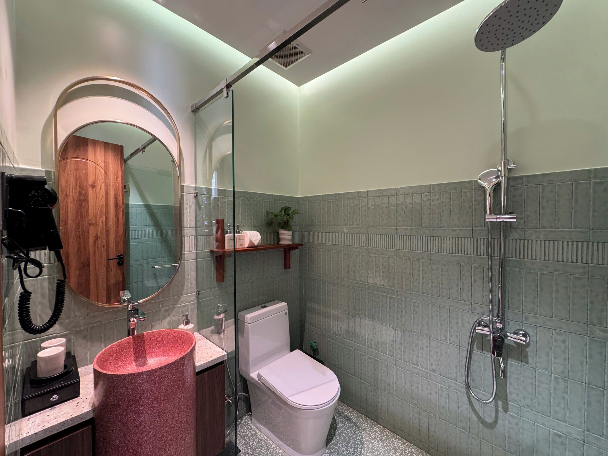 A modern bathroom with a round mirror, a pink sink, a toilet, a shower, and a wooden door.