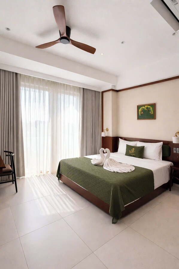Une chambre d’hôtel avec un lit, un ventilateur de plafond, une grande fenêtre avec des rideaux, une chaise et un tableau au mur.