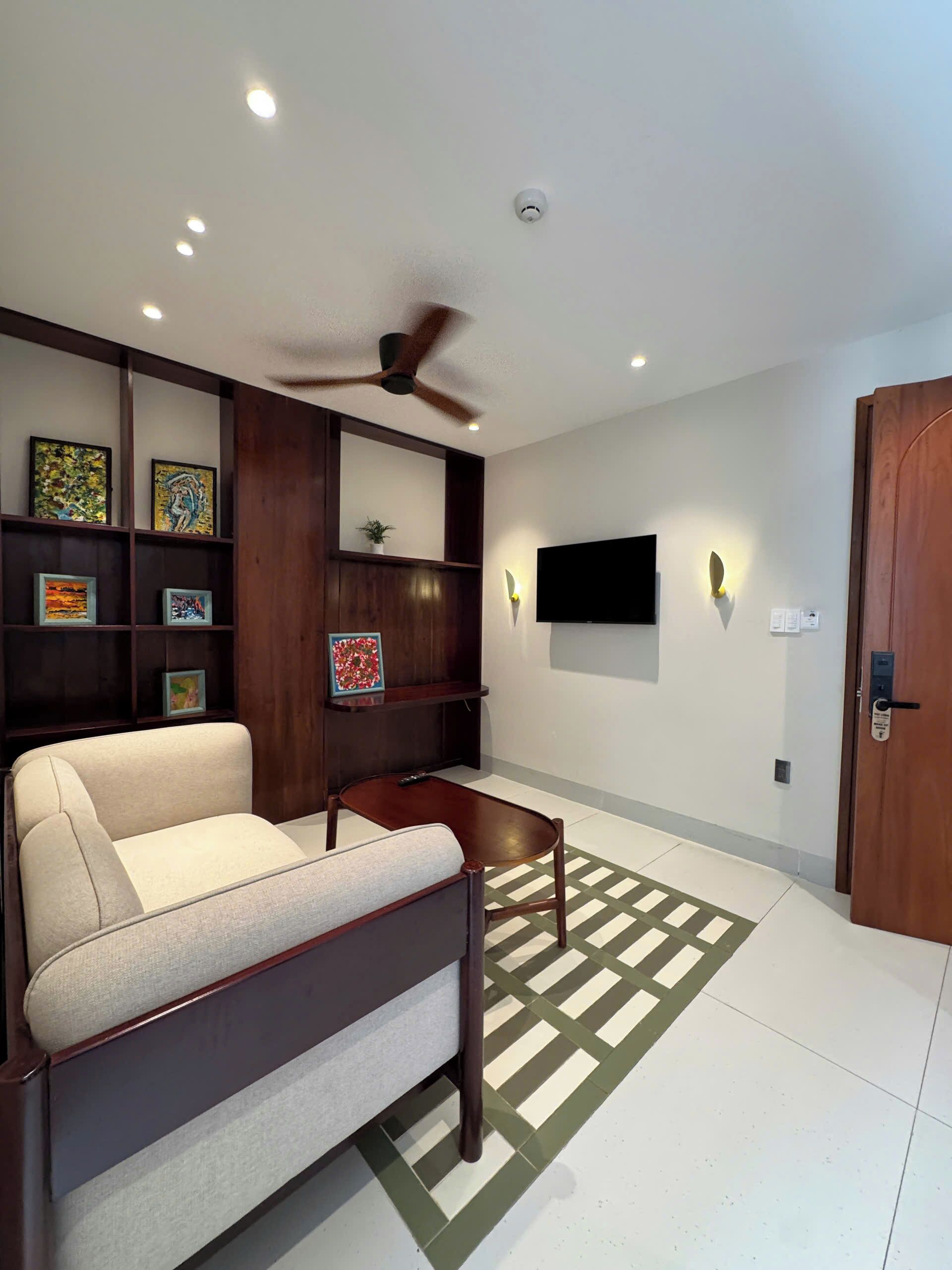 A living room with a sofa, a coffee table, a ceiling fan, wall-mounted TV, and shelves with framed artwork.