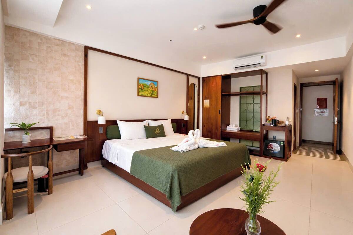 A hotel room with a bed, a desk, a chair, a vase with flowers, and a ceiling fan.