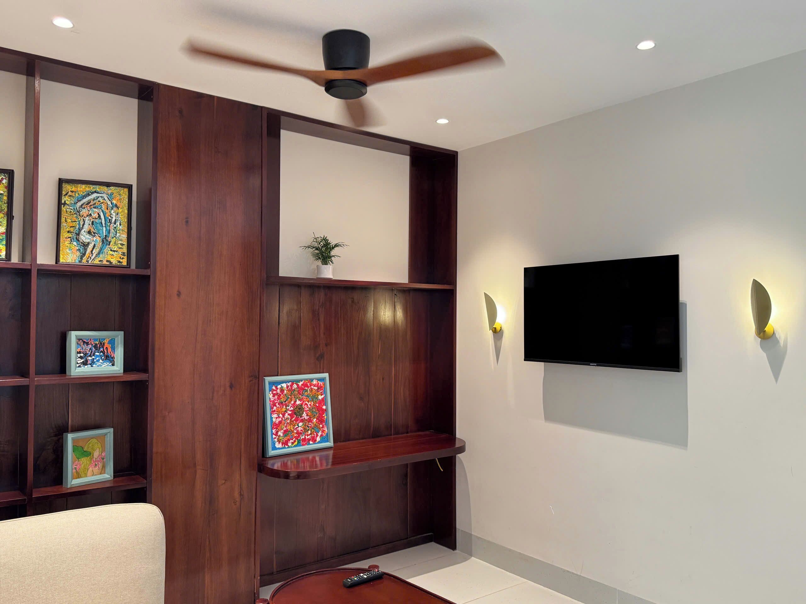 Una sala moderna con televisor montado, estanterías de madera con obras de arte, ventilador de techo e iluminación empotrada.