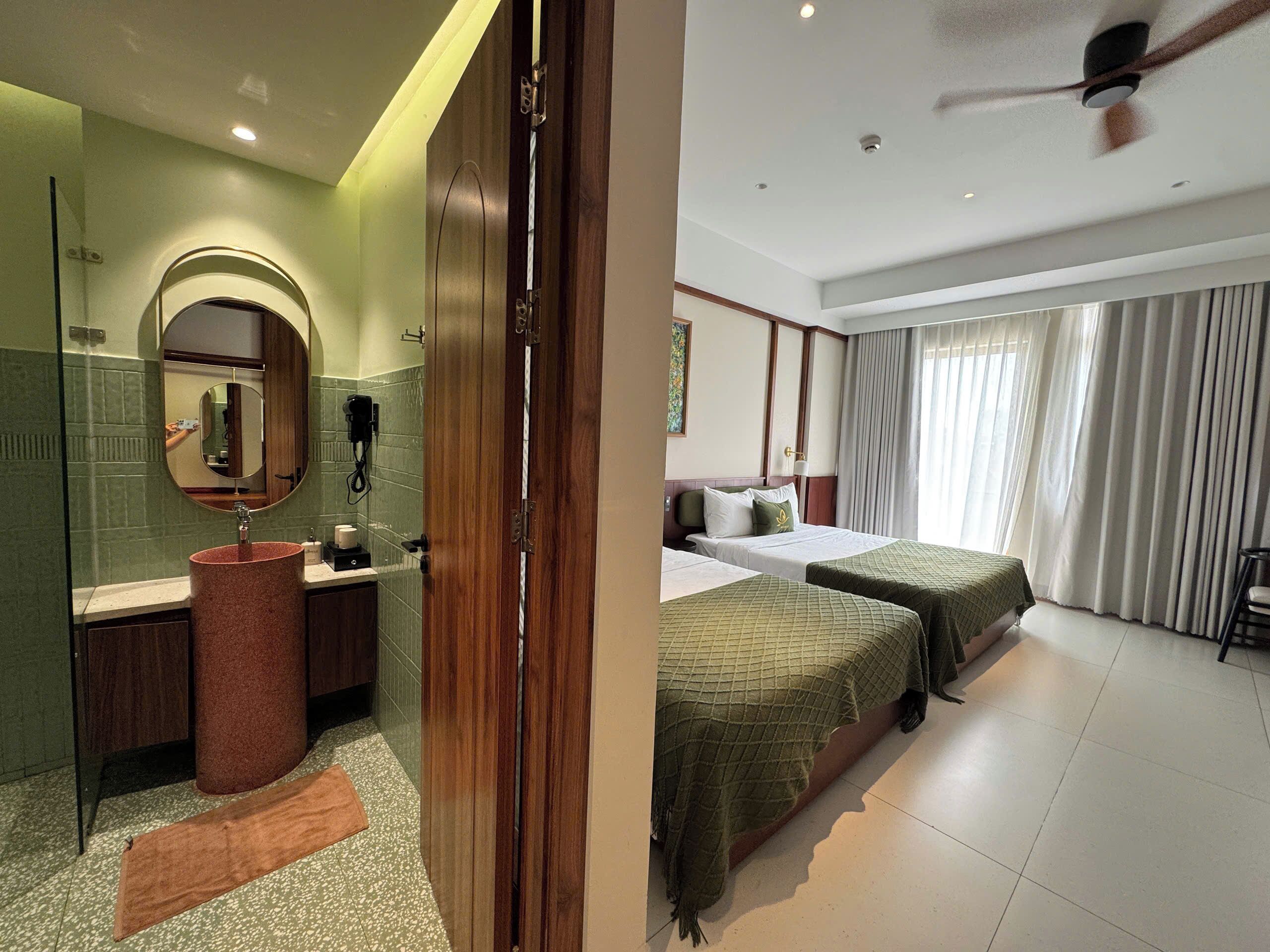 A hotel room with a bathroom, a bed with two beds, a ceiling fan, and a large window with curtains.