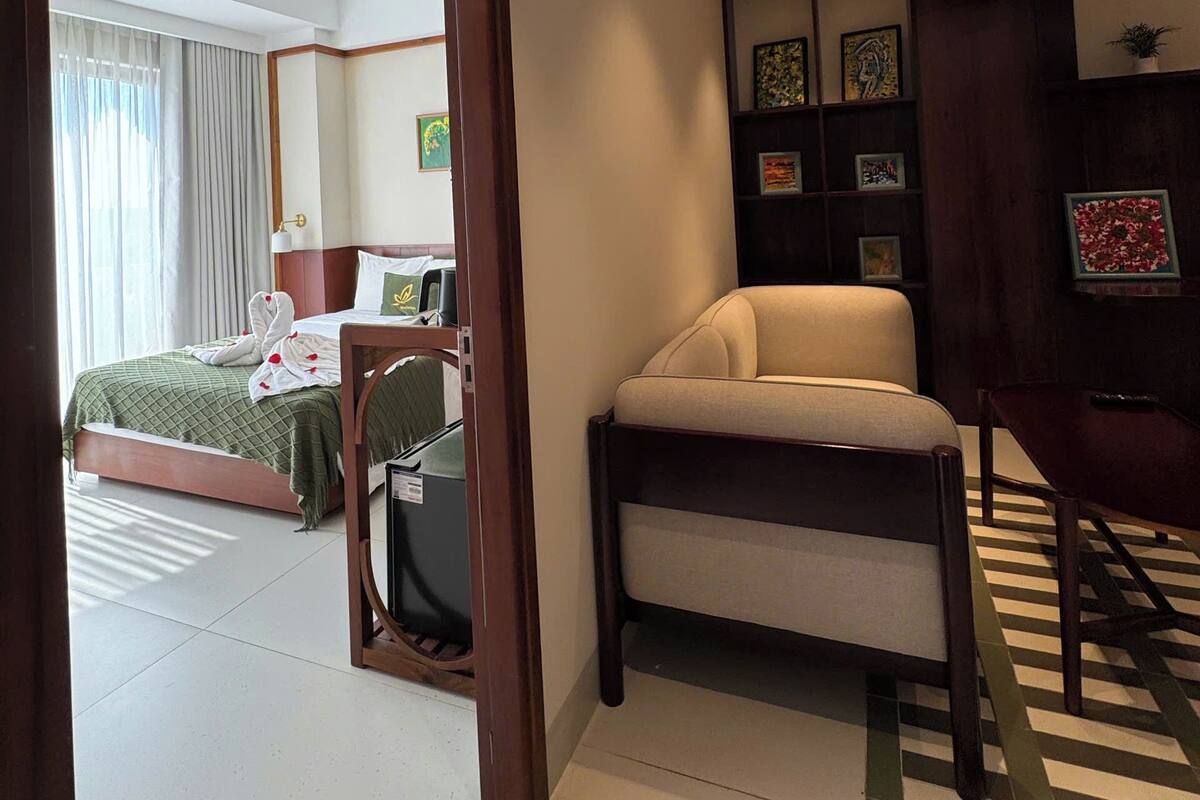 A hotel room with a bed, a desk, a chair, a bookshelf, and a ceiling fan.