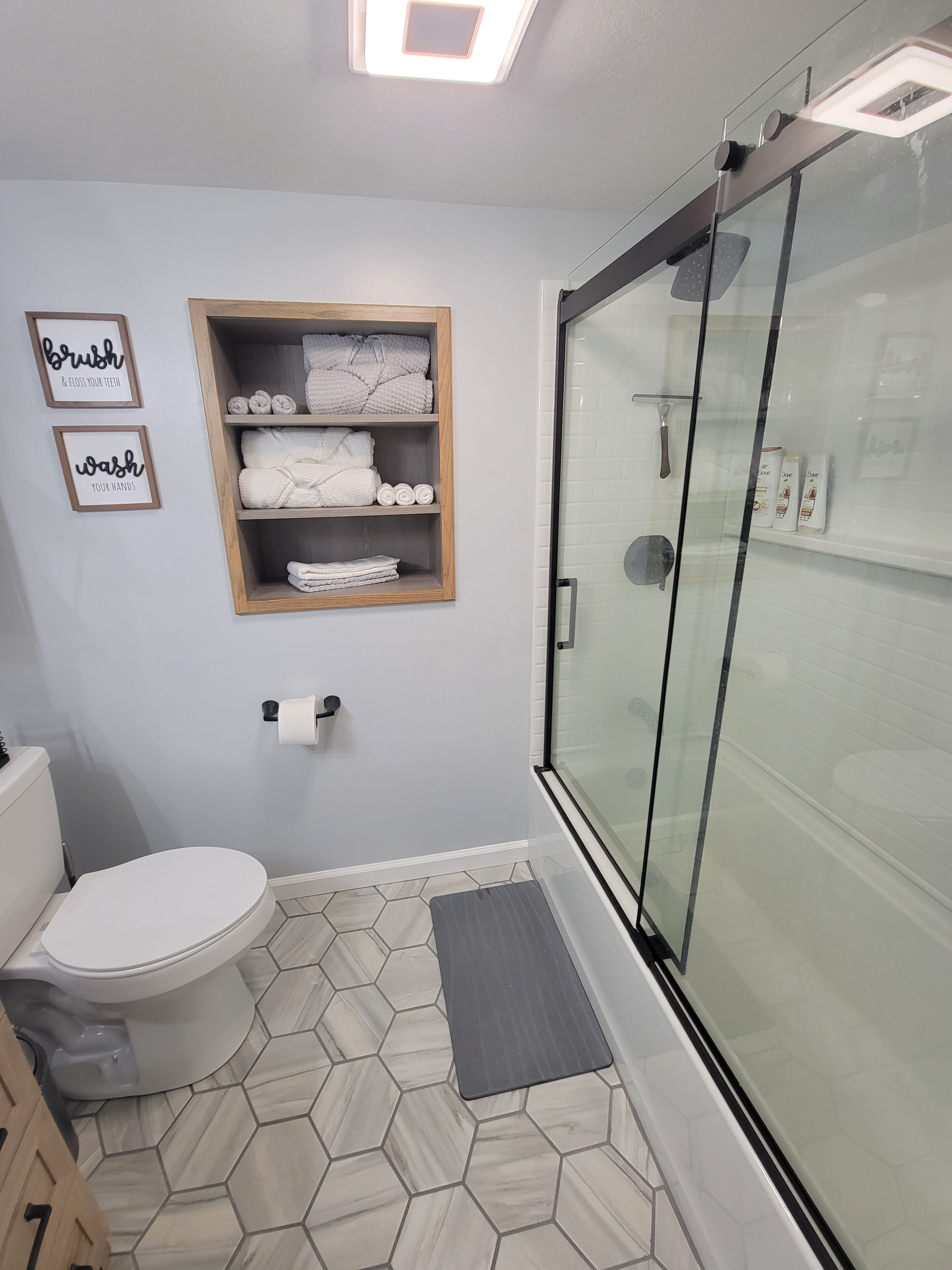 Ein modernes Badezimmer mit Toilette, einer Duschkabine aus Glas, einem Holzregal mit Handtüchern und zwei gerahmten Schildern an der Wand.