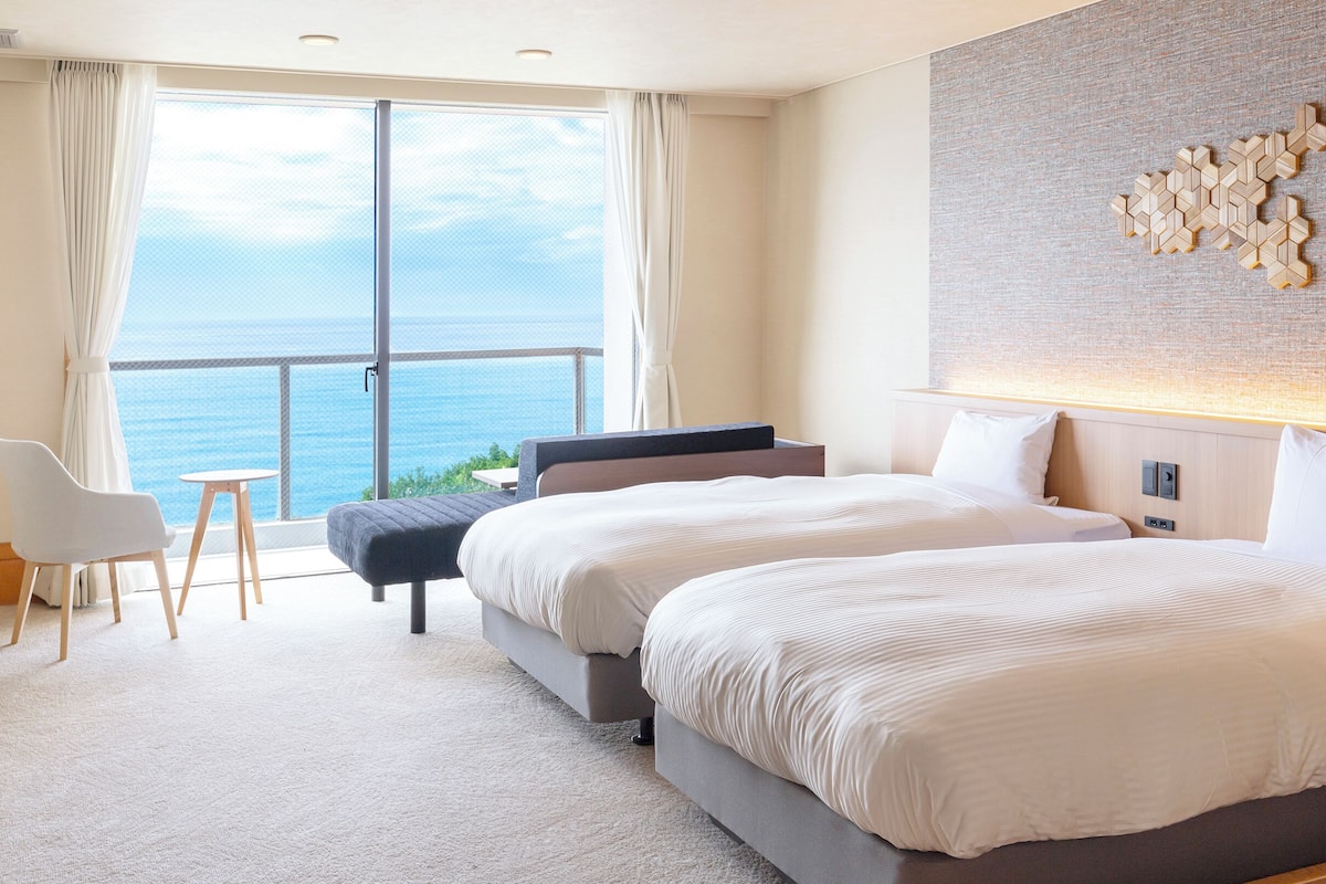 A hotel room with a large window, two beds, a desk, and a chair.