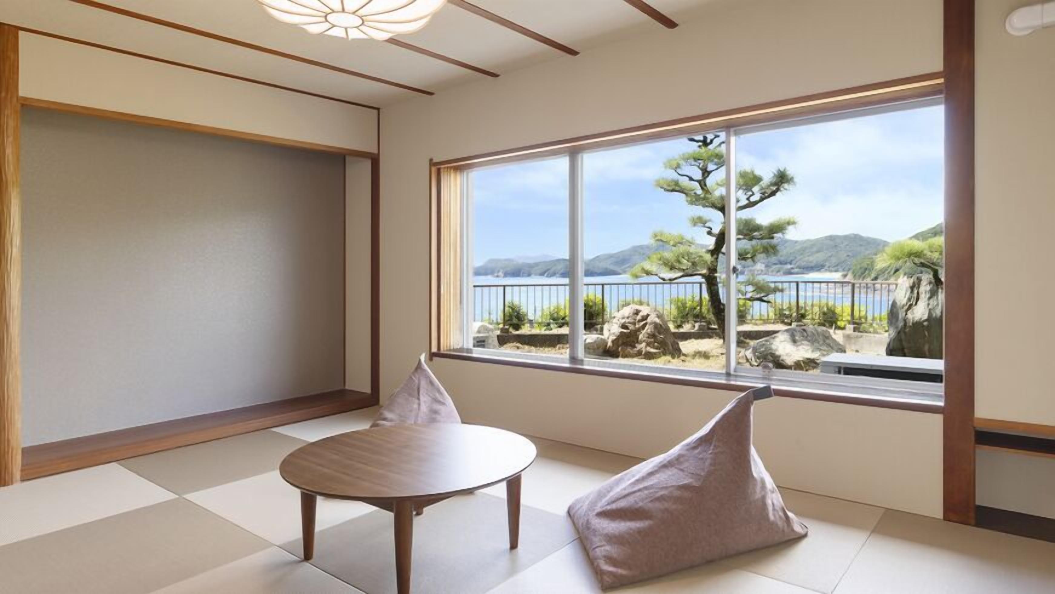 Una habitación con suelo de tatami, una mesa de madera, dos cojines y un gran ventanal con vistas a un árbol y montañas.