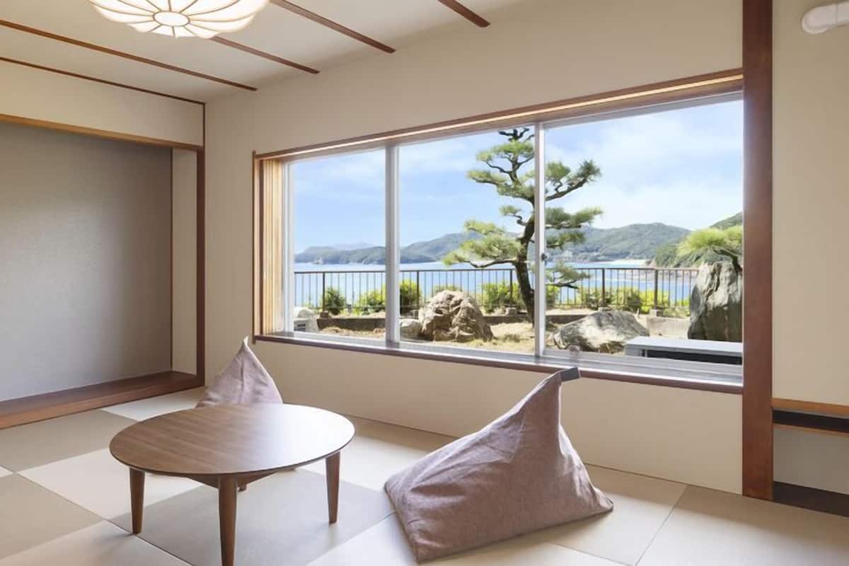 A room with tatami flooring, a wooden table, two cushions, and a large window with a view of a tree and mountains.