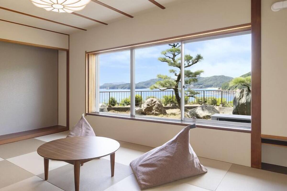 Una habitación con suelo de tatami, una mesa de madera, dos cojines y un gran ventanal con vistas a un árbol y montañas.