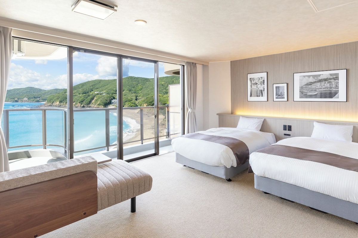 A hotel room with two beds, a large window with a view of the sea, and a balcony.