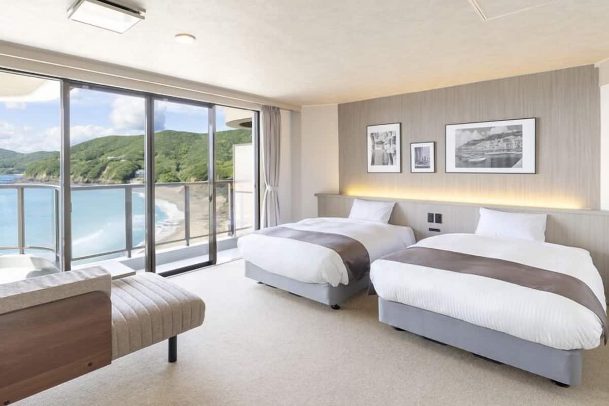 A modern hotel room with two beds, a large window with a view of the sea, and a balcony.