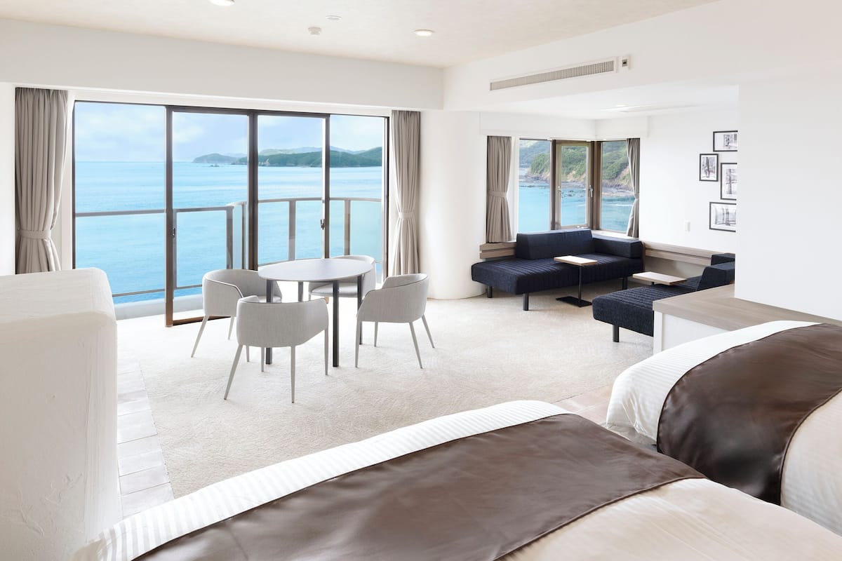 A modern hotel room with a large balcony, a sofa, a dining table, and a bed.