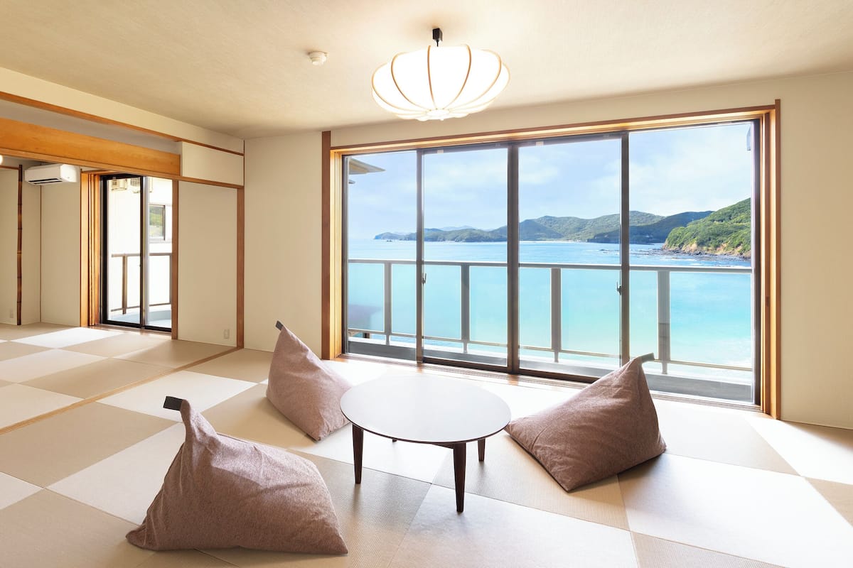 A room with tatami flooring, a sliding door, a view of the sea, and a table with cushions.