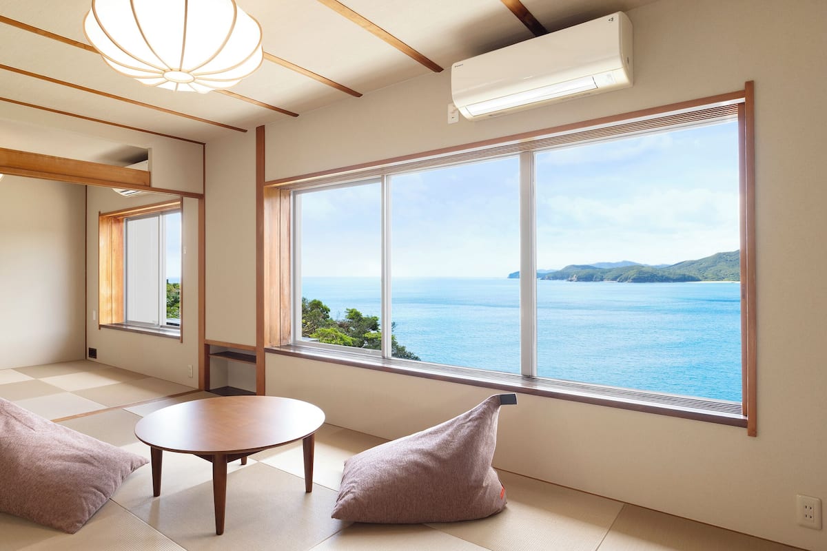 A room with tatami flooring, a wooden table, two cushions, a large window with a view of the sea, and a ceiling light fixture.