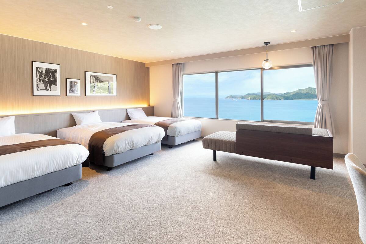 A hotel room with two beds, a seating area, and a large window with a view of the sea.