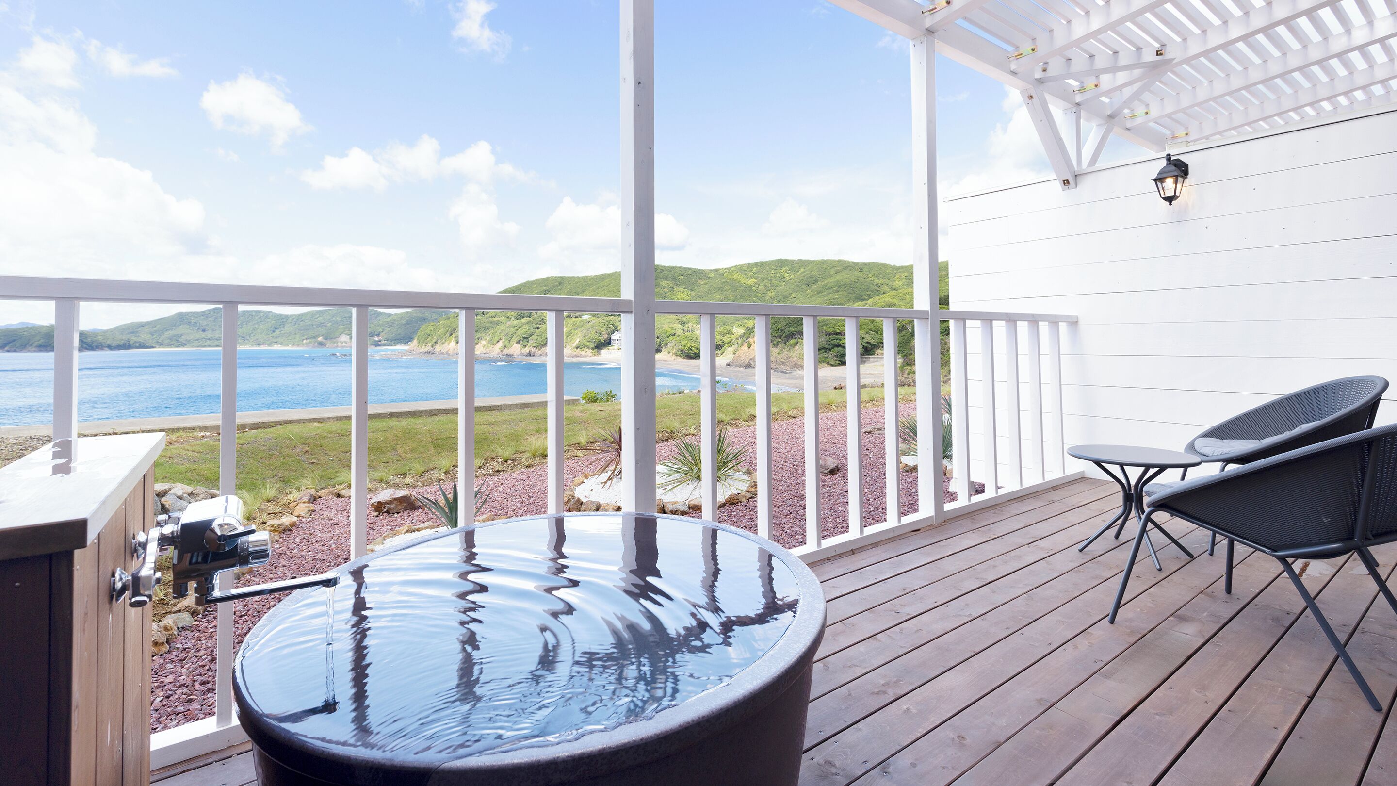A wooden deck with a hot tub, two chairs, and a view of the sea.