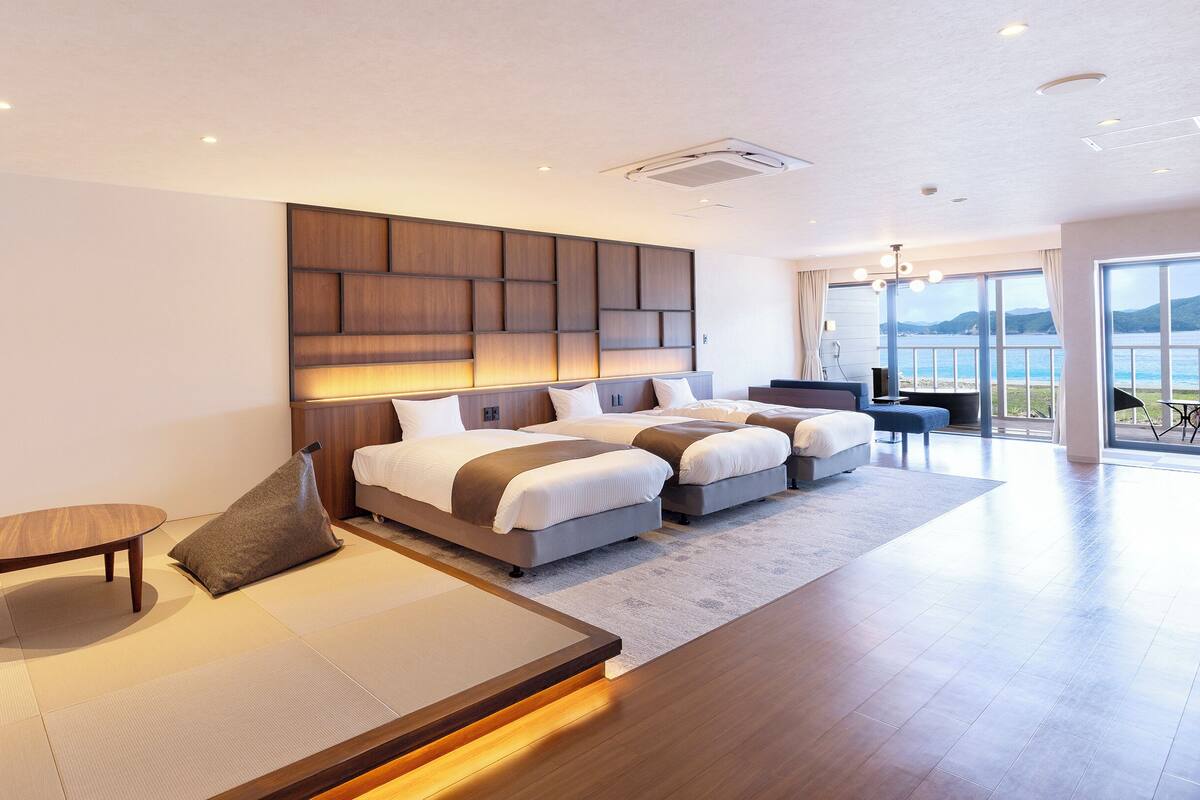 A modern hotel room with a large bed, a sofa, a small table, and a view of the sea.