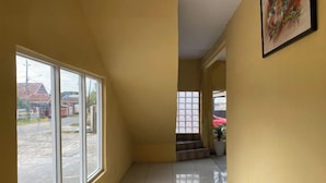 A hallway with yellow walls and a tiled floor, leading to a staircase and a window with a view of a street and buildings. - Hotel Warna (Palopo)