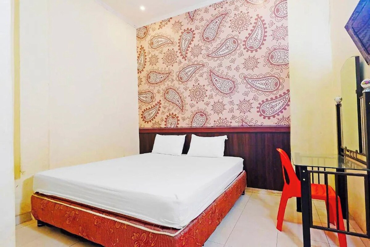 Un letto singolo con materasso bianco e testiera decorata con motivi floreali. Una sedia rossa è posizionata alla destra del letto.