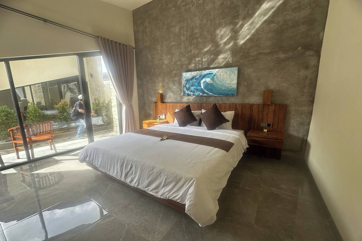 A modern hotel room with a large bed, a wooden headboard, a painting of a wave, and a balcony with a view.