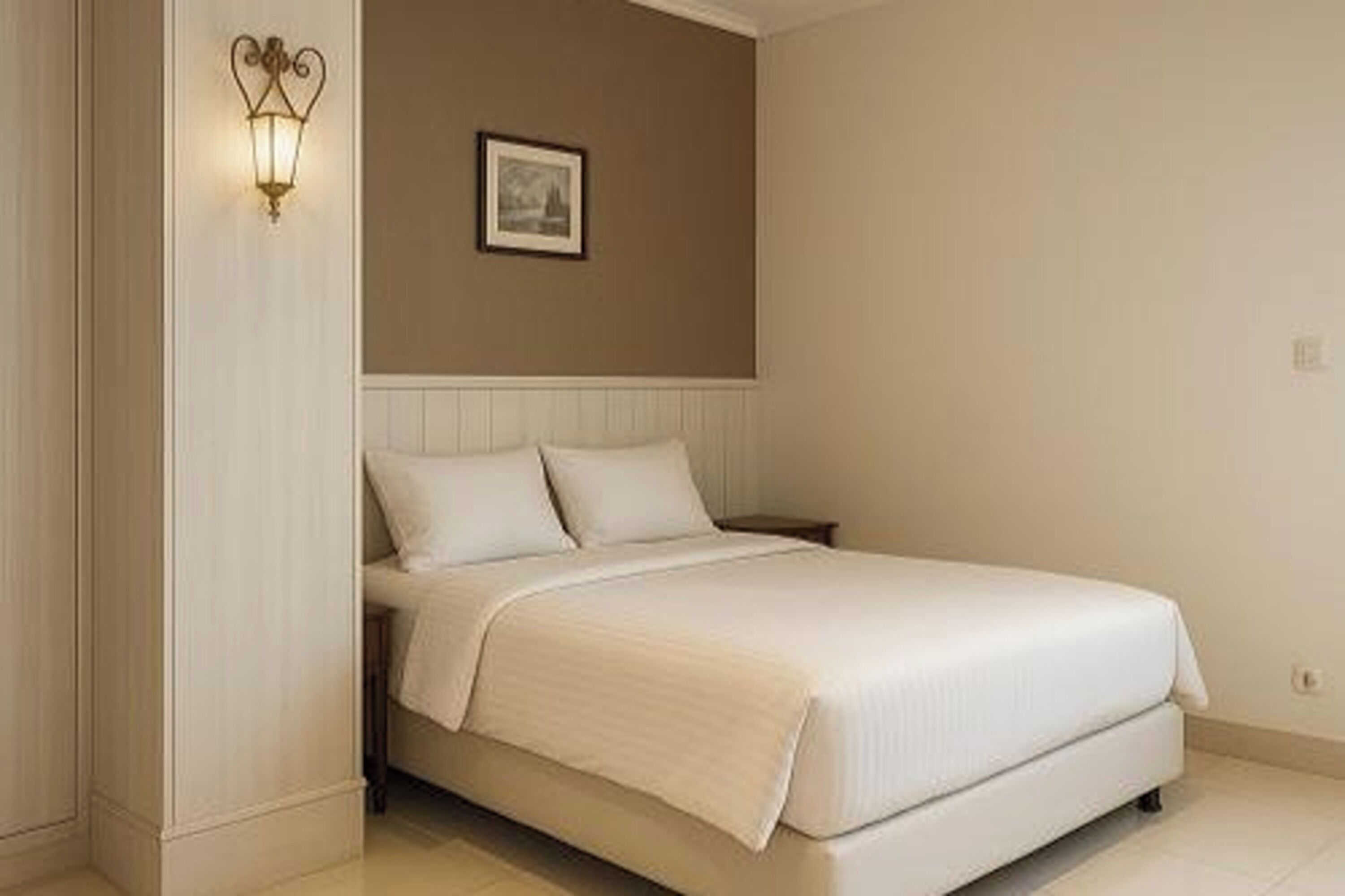 Een net opgemaakt bed met een witte deken en kussens, een wandlamp en een ingelijst foto aan de muur.
