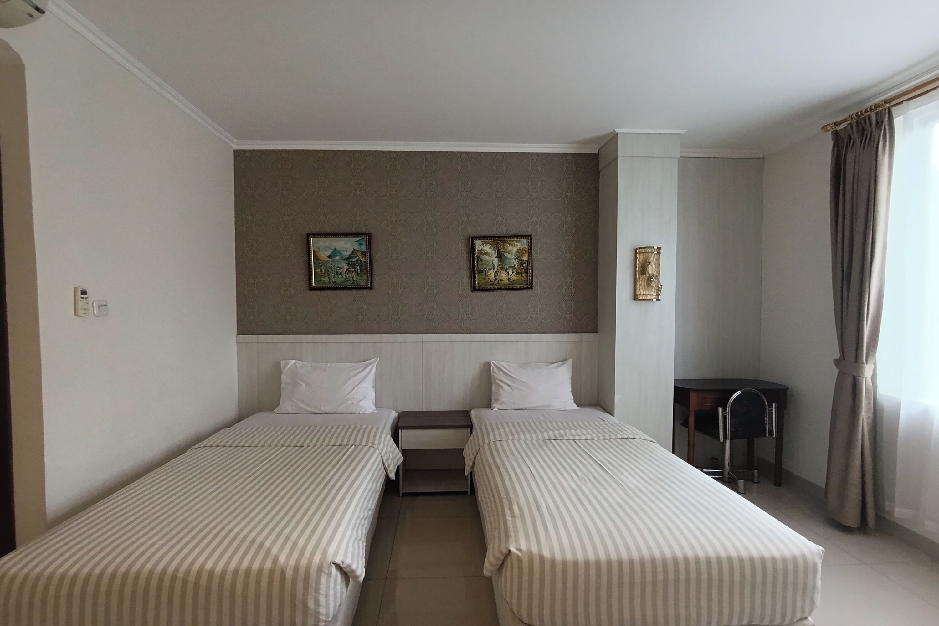 A hotel room with two single beds, a small desk, and a chair. There are two framed pictures on the wall above the beds.