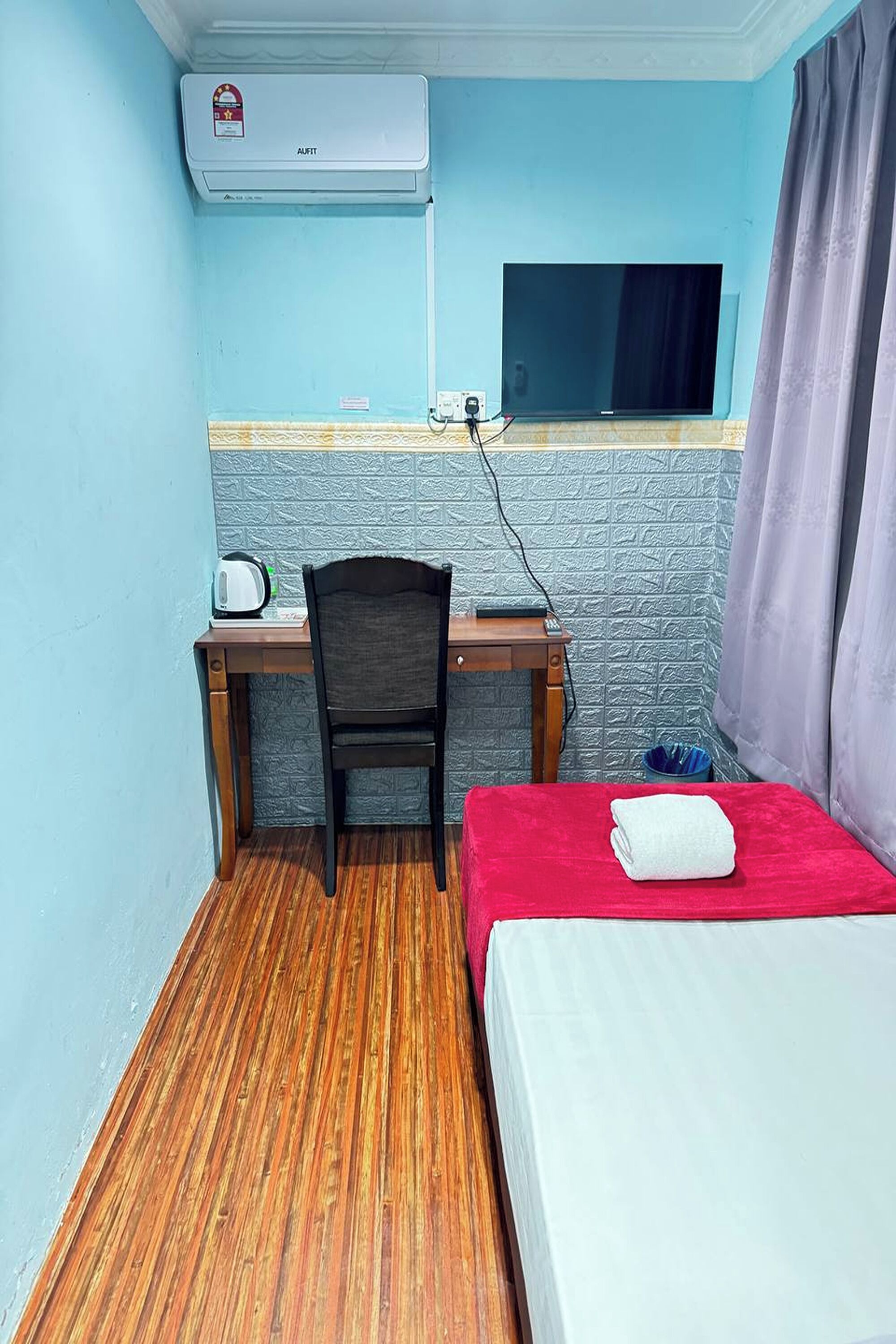 Una habitación pequeña con un escritorio de madera, una silla, una cama con cubierta roja, un televisor montado en la pared y un aire acondicionado.
