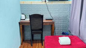 Una habitación pequeña con un escritorio de madera, una silla, una cama con cubierta roja, un televisor montado en la pared y un aire acondicionado.