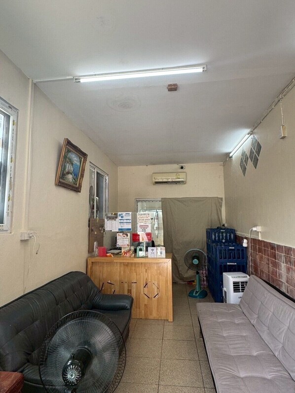 A small, compact room with a wooden desk, a black leather couch, a grey sofa, and a standing fan. - Sukhum Suvarnabhumi Airport Guest House (Bangkok)