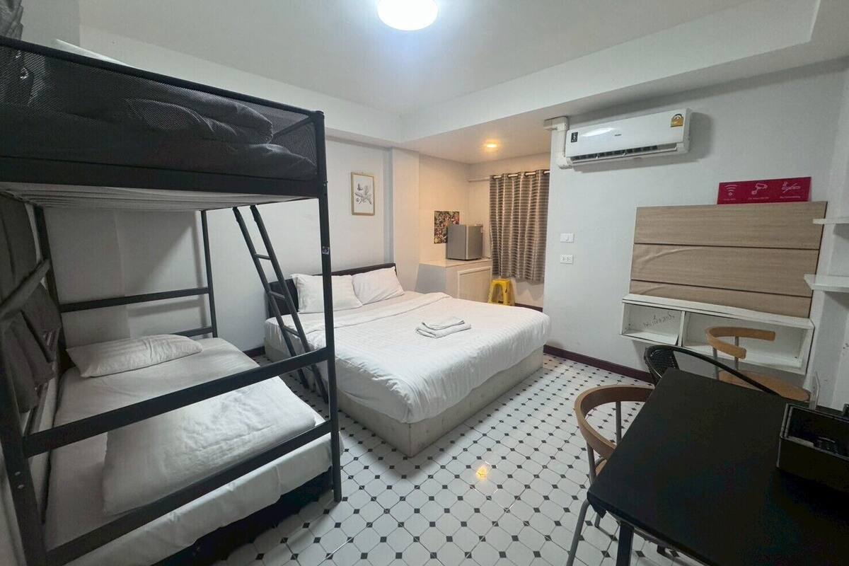 A room with bunk beds, a desk, a chair, an air conditioning unit, and a small table with a lamp.
