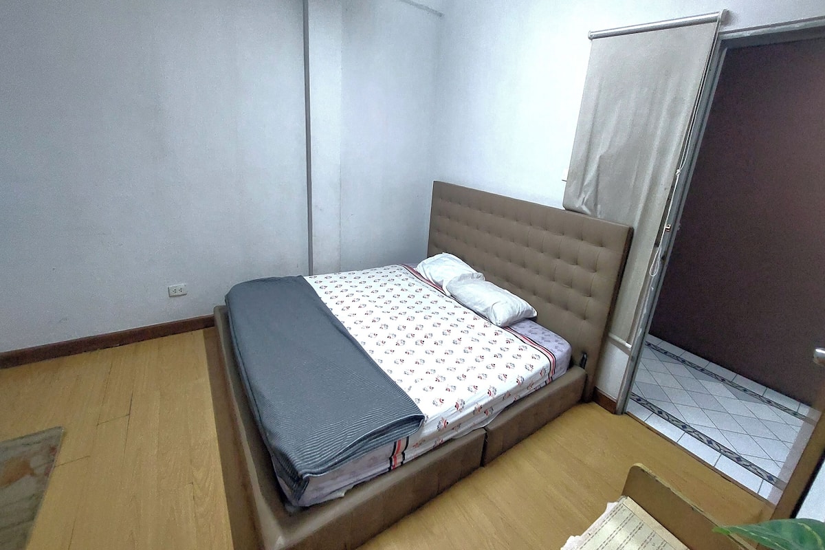 Une petite chambre avec un lit simple, un plancher en bois, une tête de lit beige et une fenêtre avec un rideau.