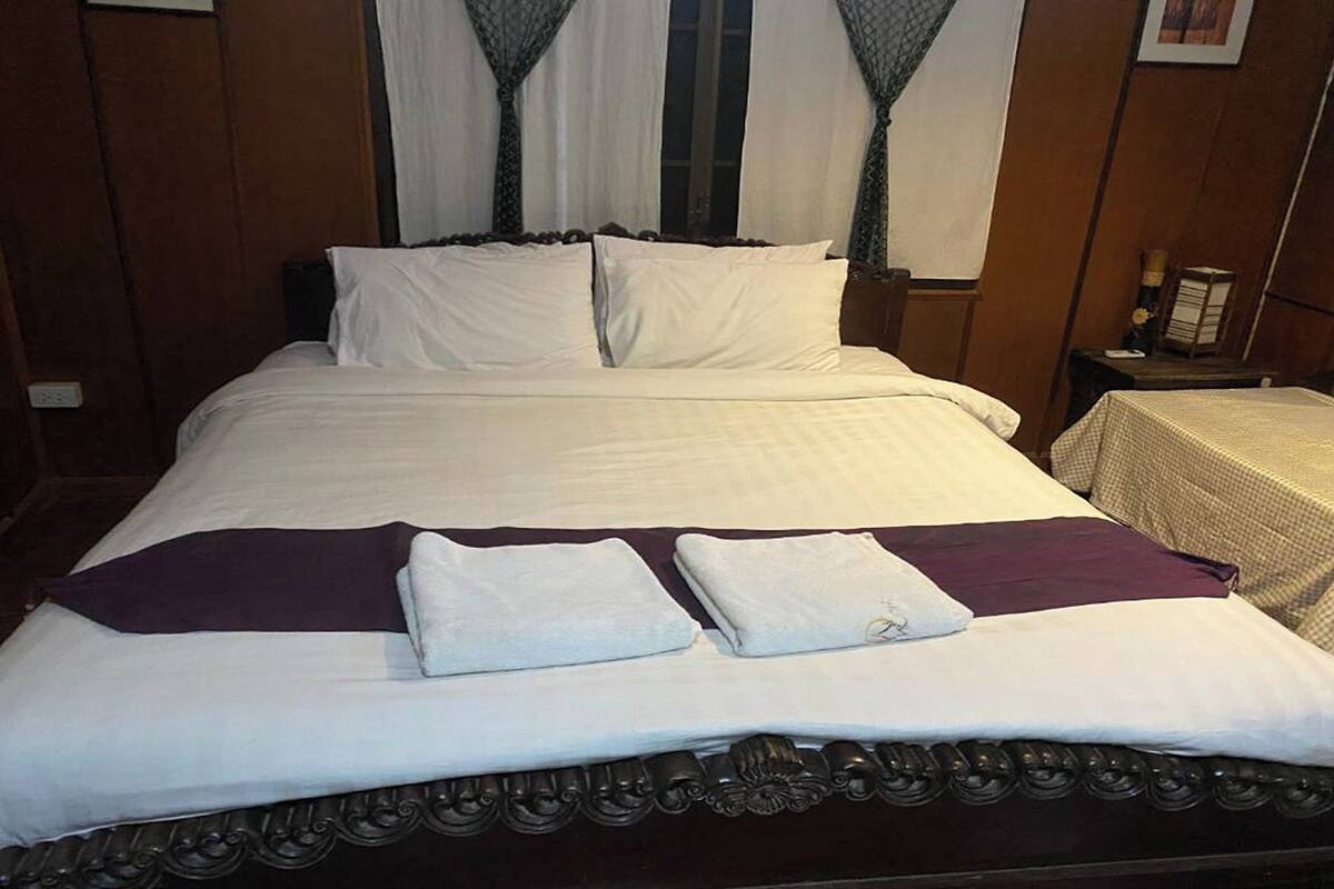 Un lit bien fait, avec du linge de lit blanc et un plaid violet, une tête de lit en bois et une petite table avec une lampe placée à côté du lit.