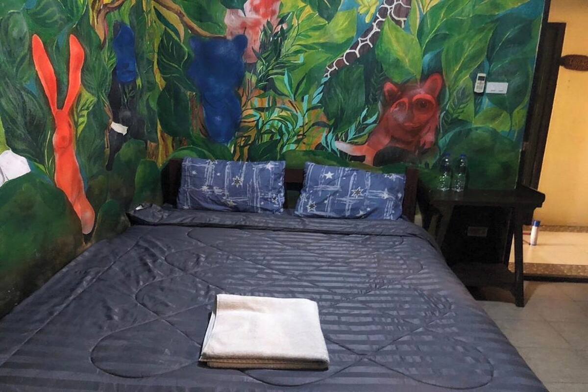 Une pièce avec un lit, une fresque murale et une petite table sur laquelle sont posées des bouteilles.
