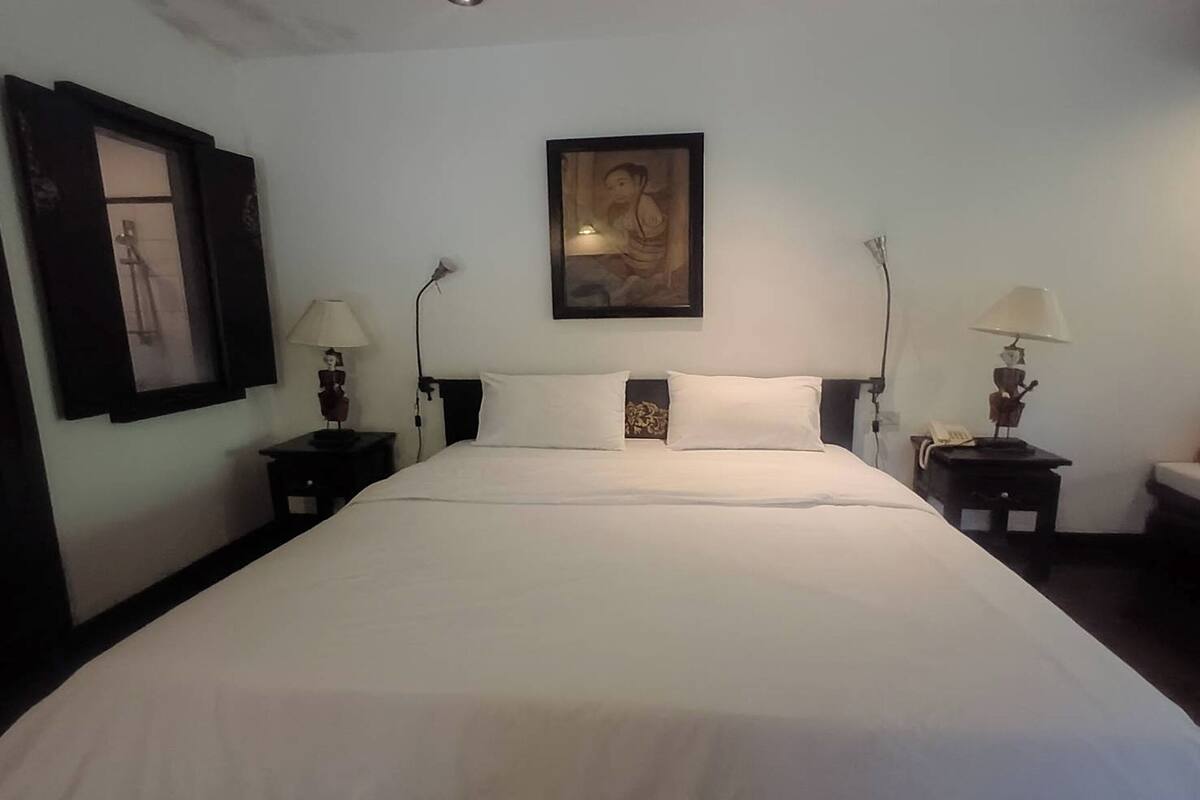 Una cama bien hecha con sábanas blancas, dos mesitas de noche con lámparas, un cuadro en la pared y un pequeño adorno sobre la cama.