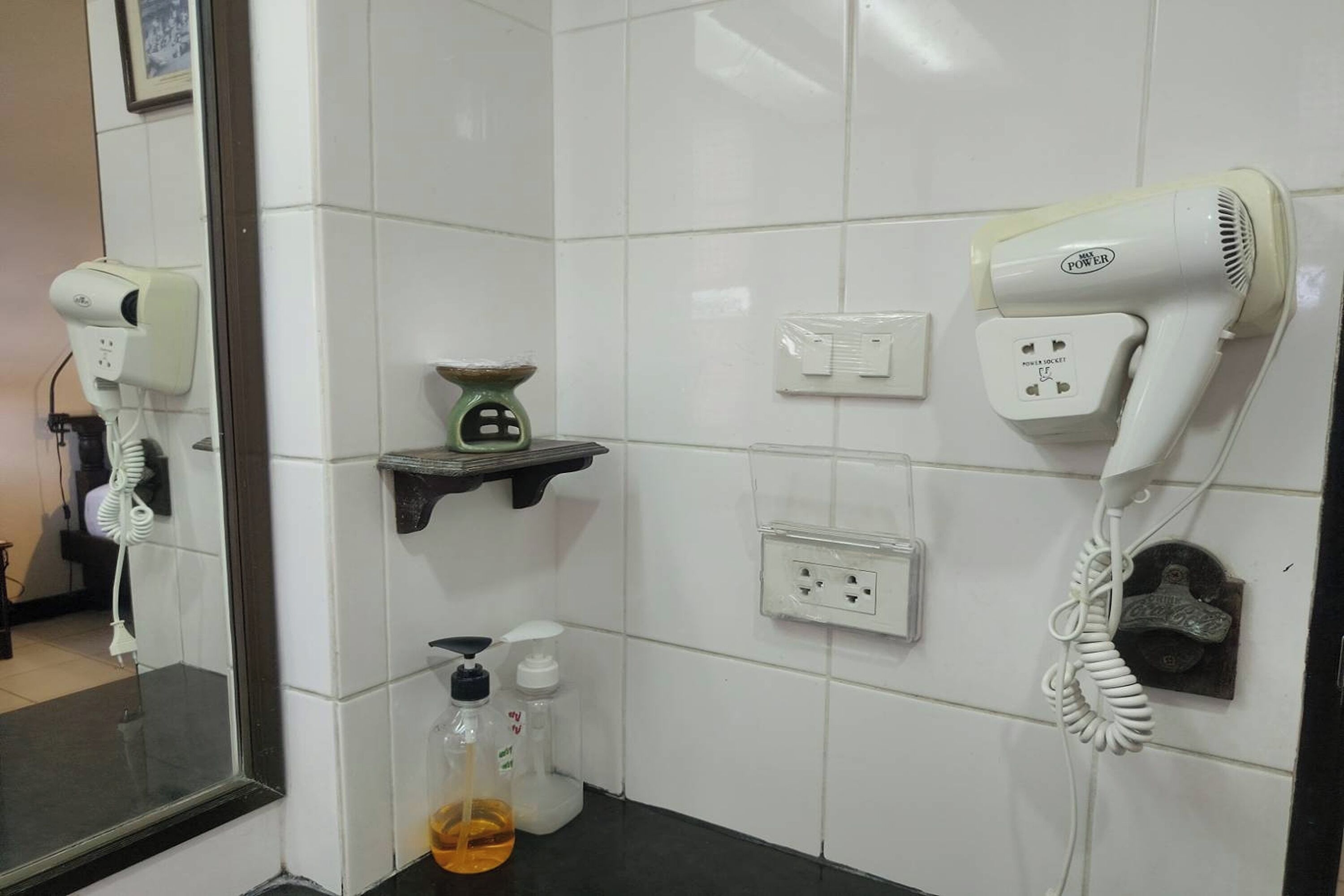Ett badrum med vita kakelplattor, en hårtork, en handtork, en tvåldispenser och en liten hylla.