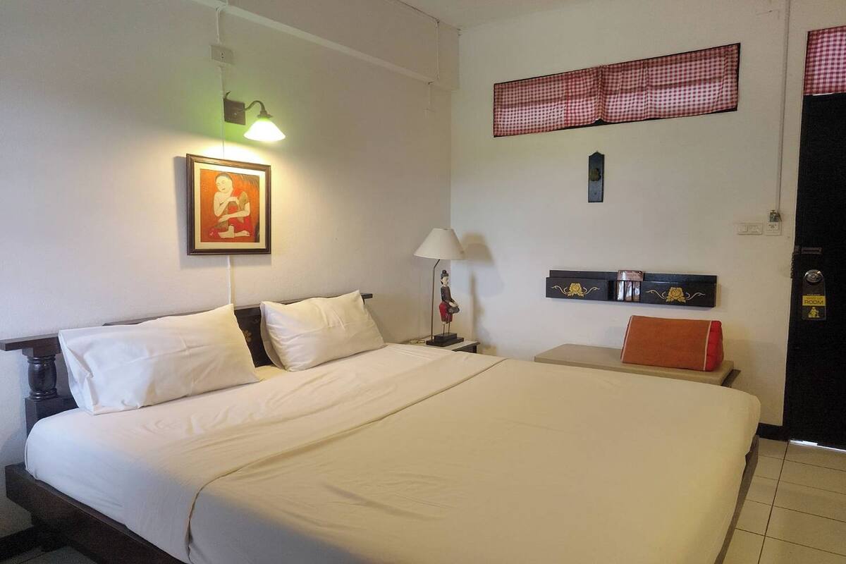 Una habitación de hotel con una cama grande, una mesita de noche con lámpara, un cuadro enmarcado en la pared y una cortina a cuadros sobre la ventana.
