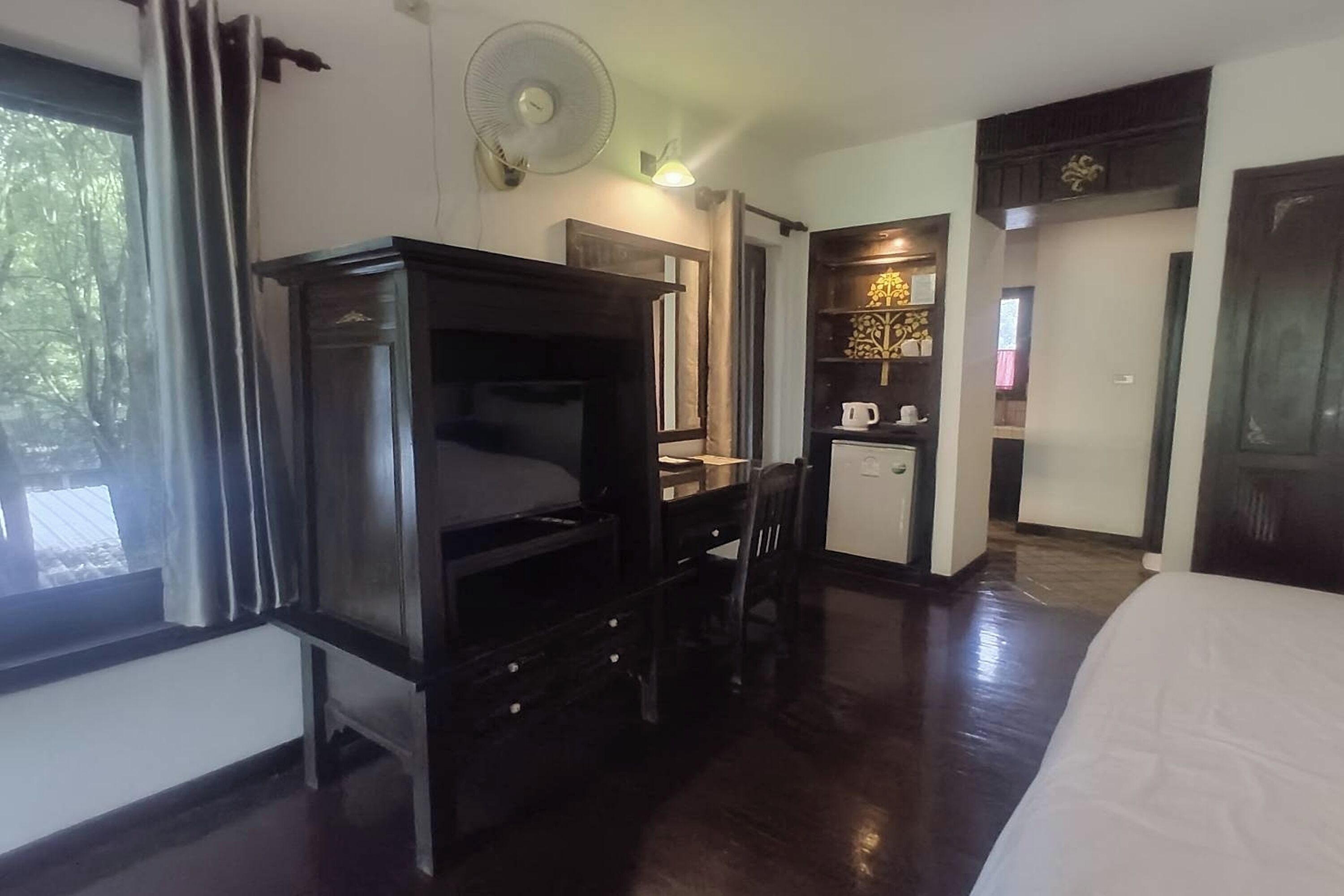 A hotel room with a bed, a desk with a chair, a wardrobe, a small kitchenette, and a window with curtains.