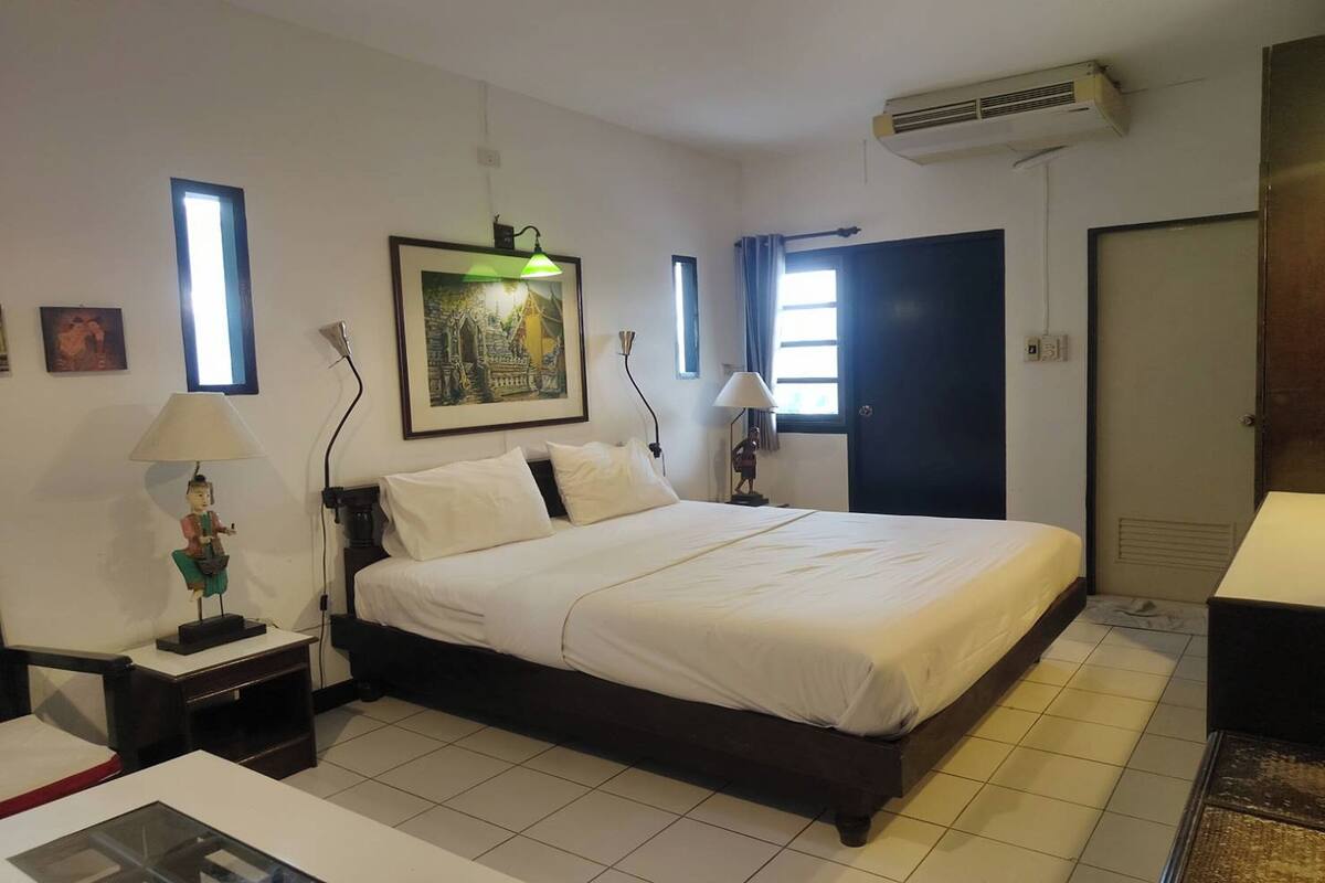 Habitación de hotel con una cama grande, dos mesitas de noche, un escritorio, una silla, una ventana con cortinas, un aire acondicionado instalado en la pared y un cuadro enmarcado.