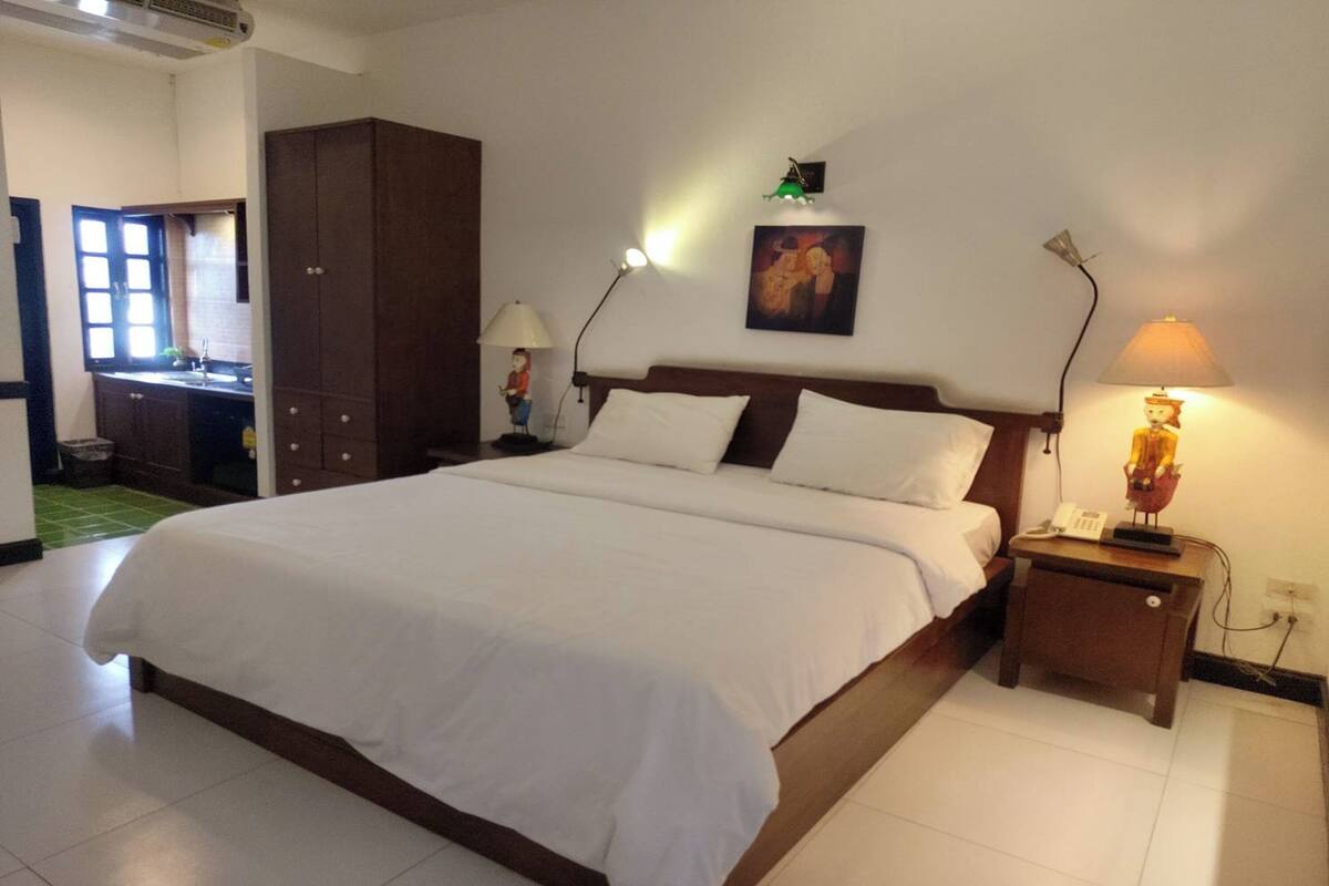 Habitación de hotel con una cama grande, una mesita de noche con lámpara, un cuadro en la pared y vistas al área de la cocina.