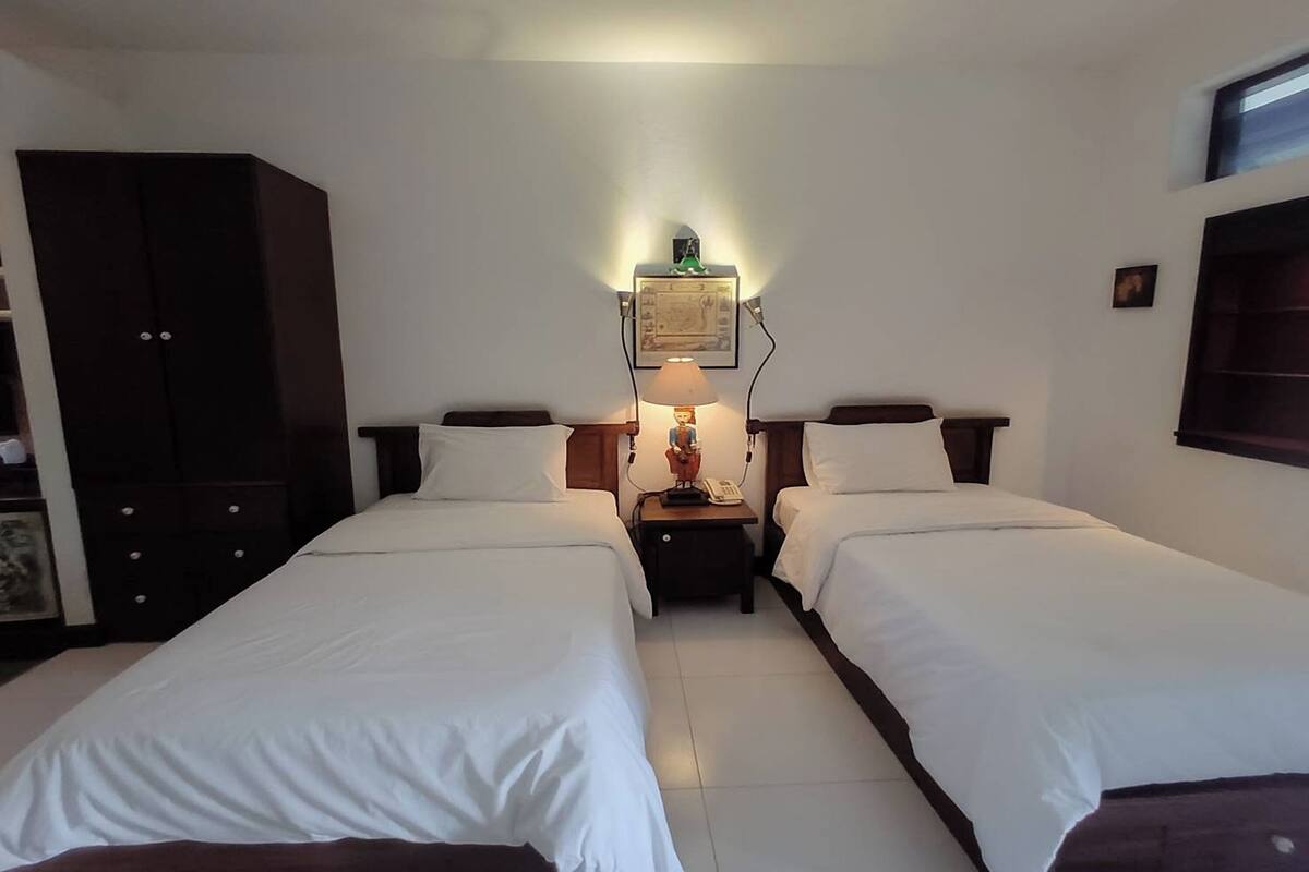 Habitación de hotel con dos camas individuales, un armario, una mesita de noche con lámpara y una lámpara de pared.