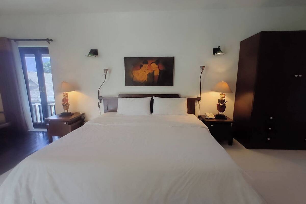 Una habitación de hotel con una cama grande, dos mesitas de noche con lámparas, un armario oscuro y un cuadro en la pared.